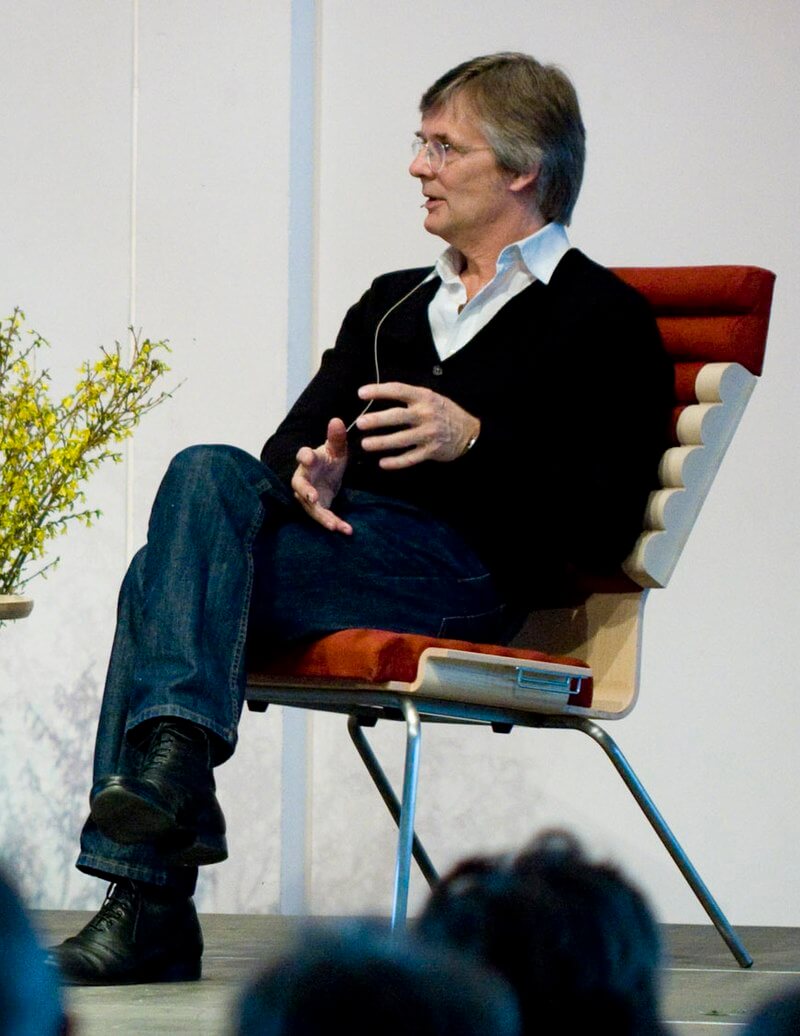 Seated on a modern chair with his legs crossed, Bille August gestures with his hands while speaking before a seated audience.