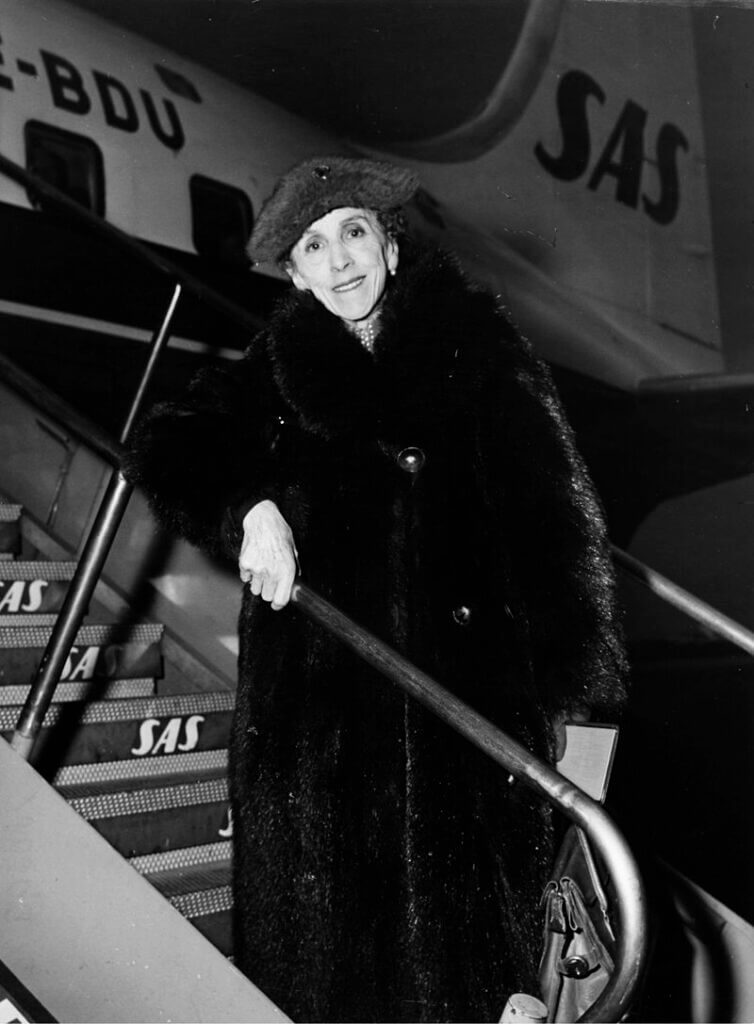 Karen Blixen, dressed in a long fur coat and complementary hat, leans on the railing of a staircase as she boards an SAS plane.