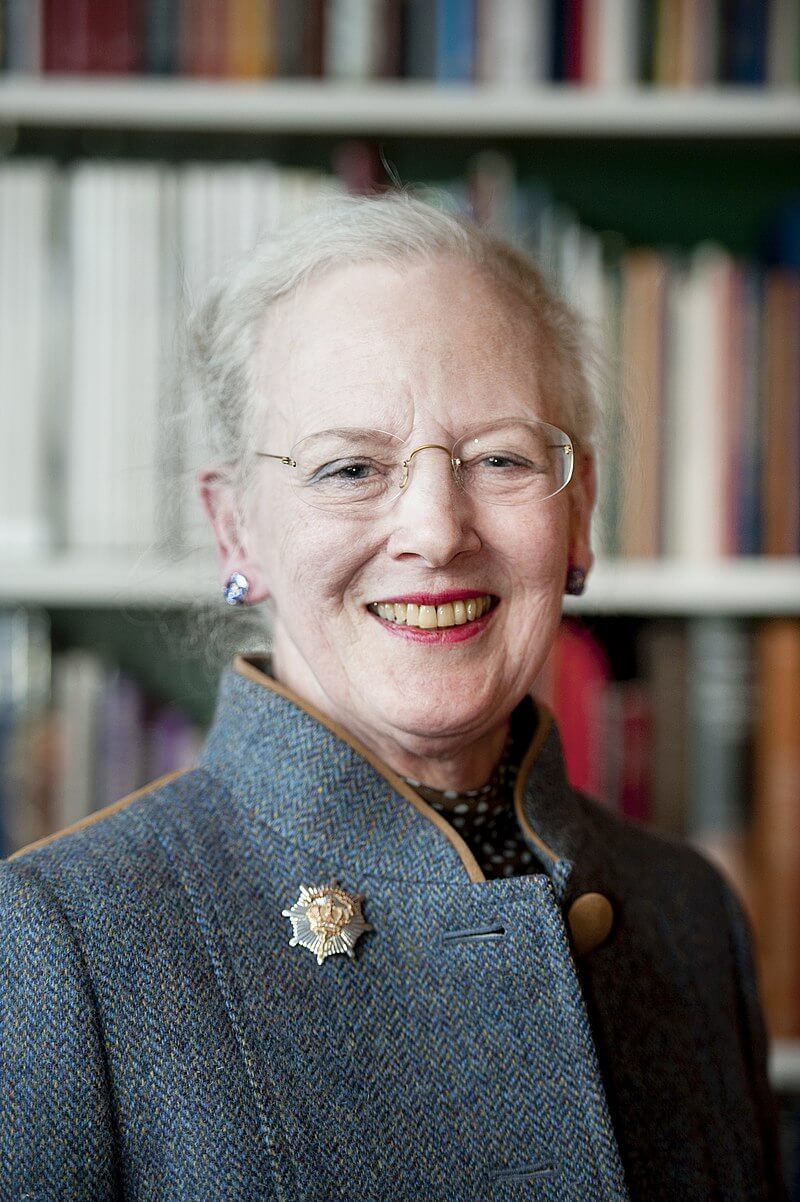Queen Margrethe dressed in a gray coat posing before a bookshelf.