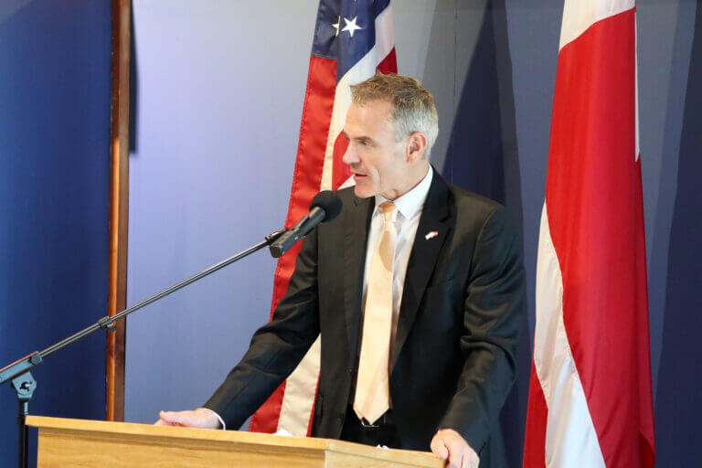 Ambassador of Denmark to the United States Jesper Møller Sørensen stands behind a podium. Danish and American flags are displayed in the background.