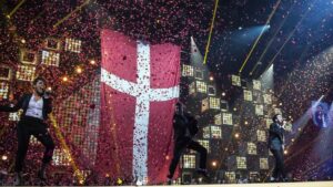 A concert with three singers, red confetti is flying around, a Danish flag is in the background of the stage.