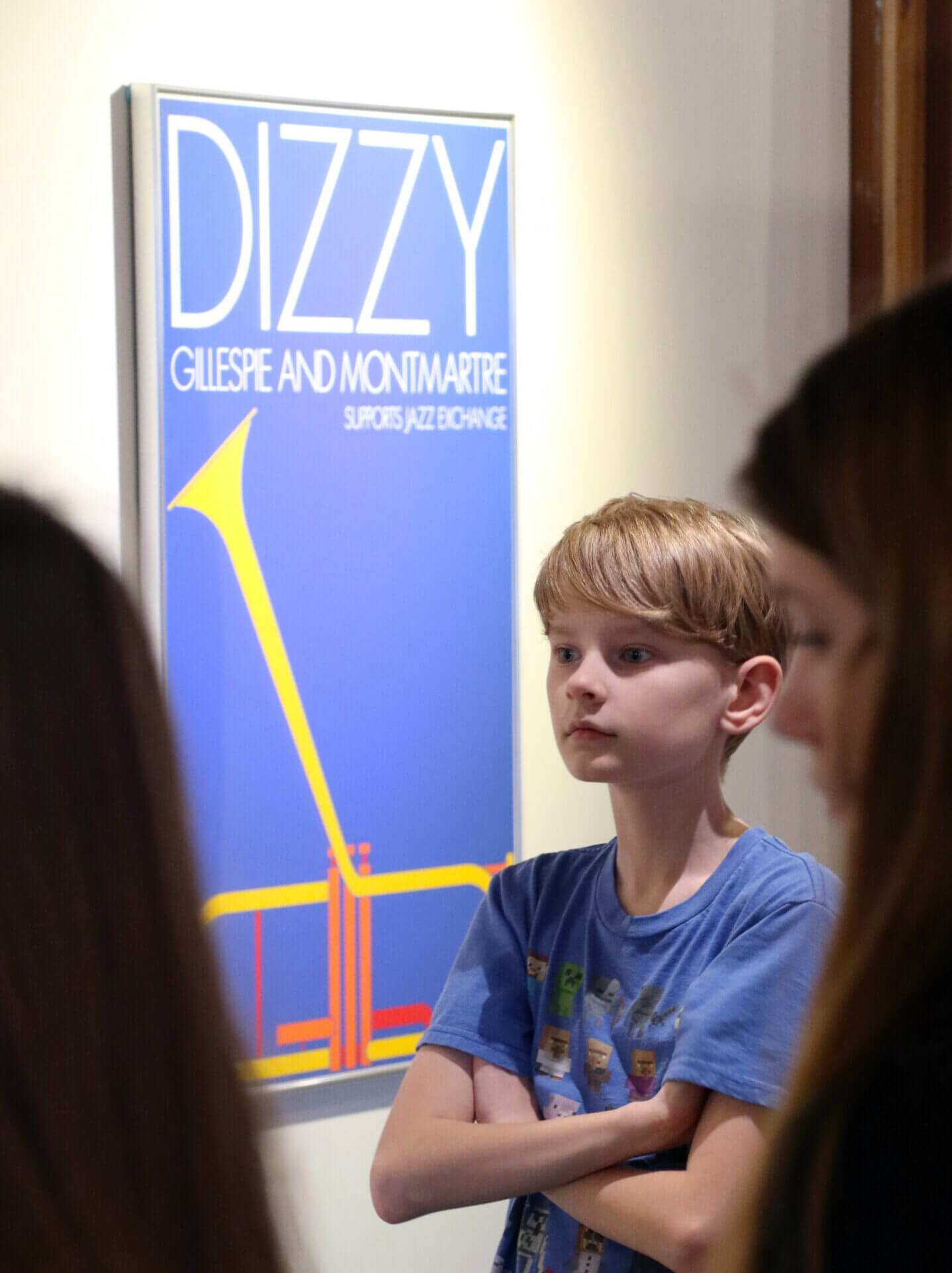 A young boy with his arms crossed stands in front of a Per Arnoldi poster during a field trip.