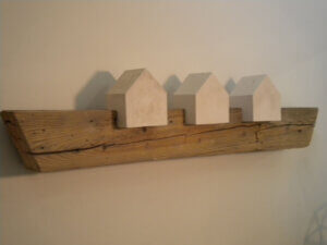 A wooden plank hangs on a wall with 3 white blocks shaped as houses imbedded within the wood.