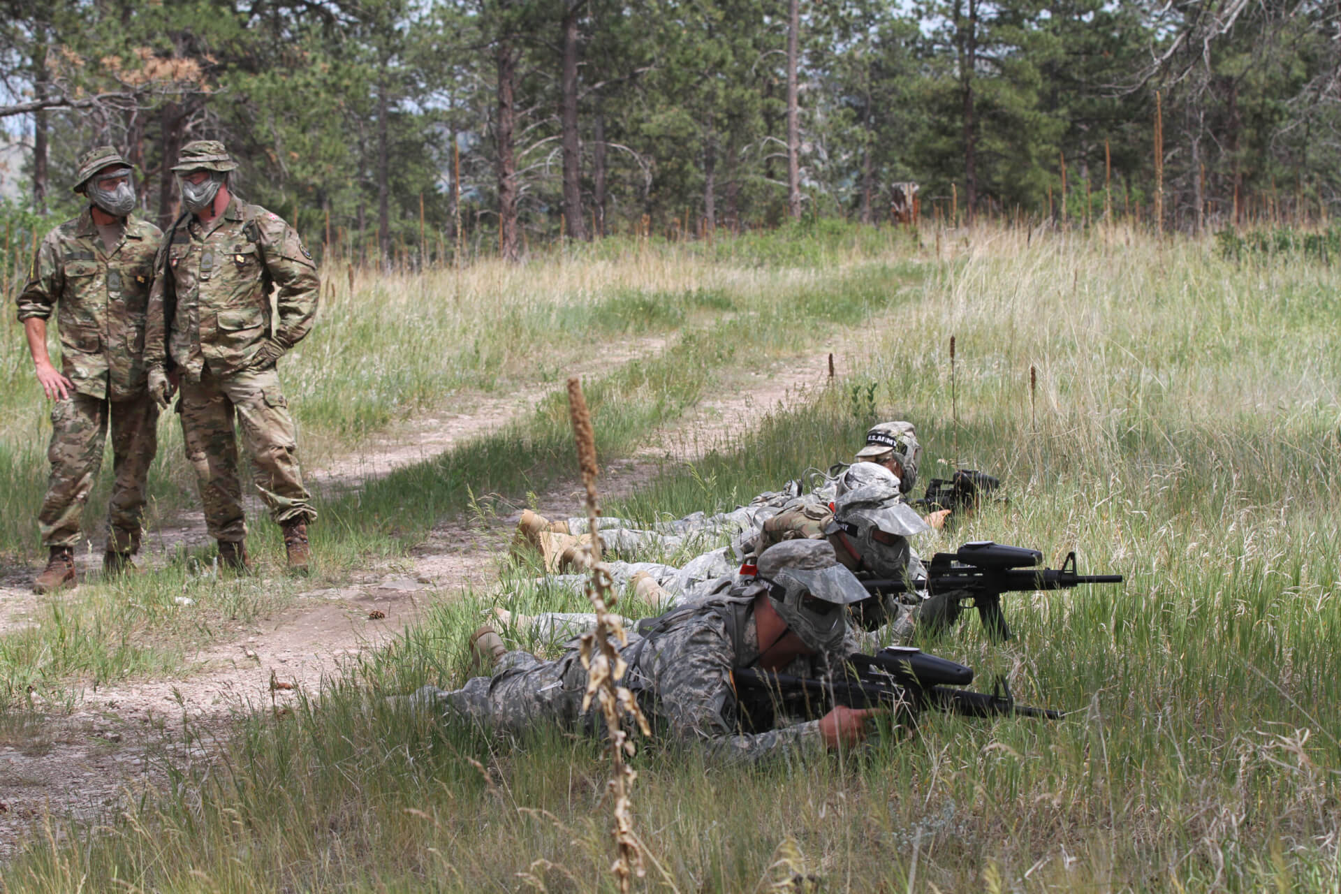 Four men in military clothes and protective masks are laying in grass, they are holding guns, behind them stands two men also in military clothing and protective masks, they are talking, in the background are trees.
