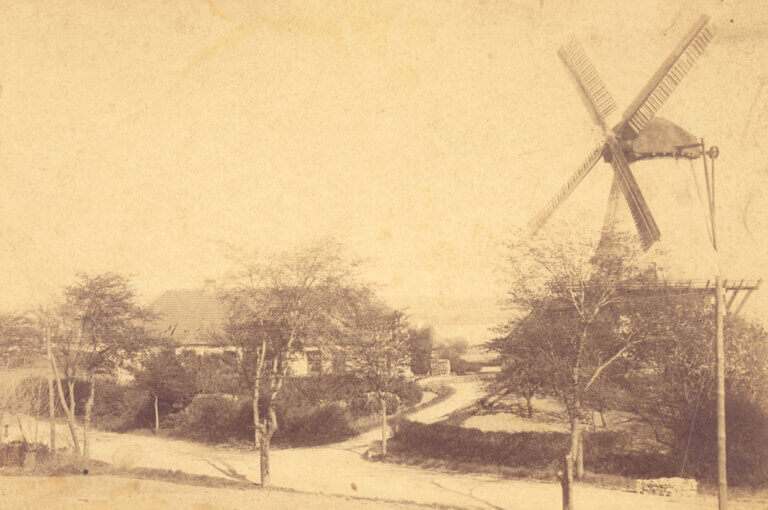 An old photograph, a windmill is on the right, a house is to the left behind some trees, in the foreground is a road.