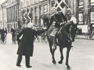 A man on a horse is shaking hands with another man on foot, behind are several men on bicycles following the man on the horse.