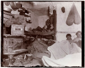 An old photography, on the left is a stove with several things stacked on and around it, in the back and to the right are several men with blankets.
