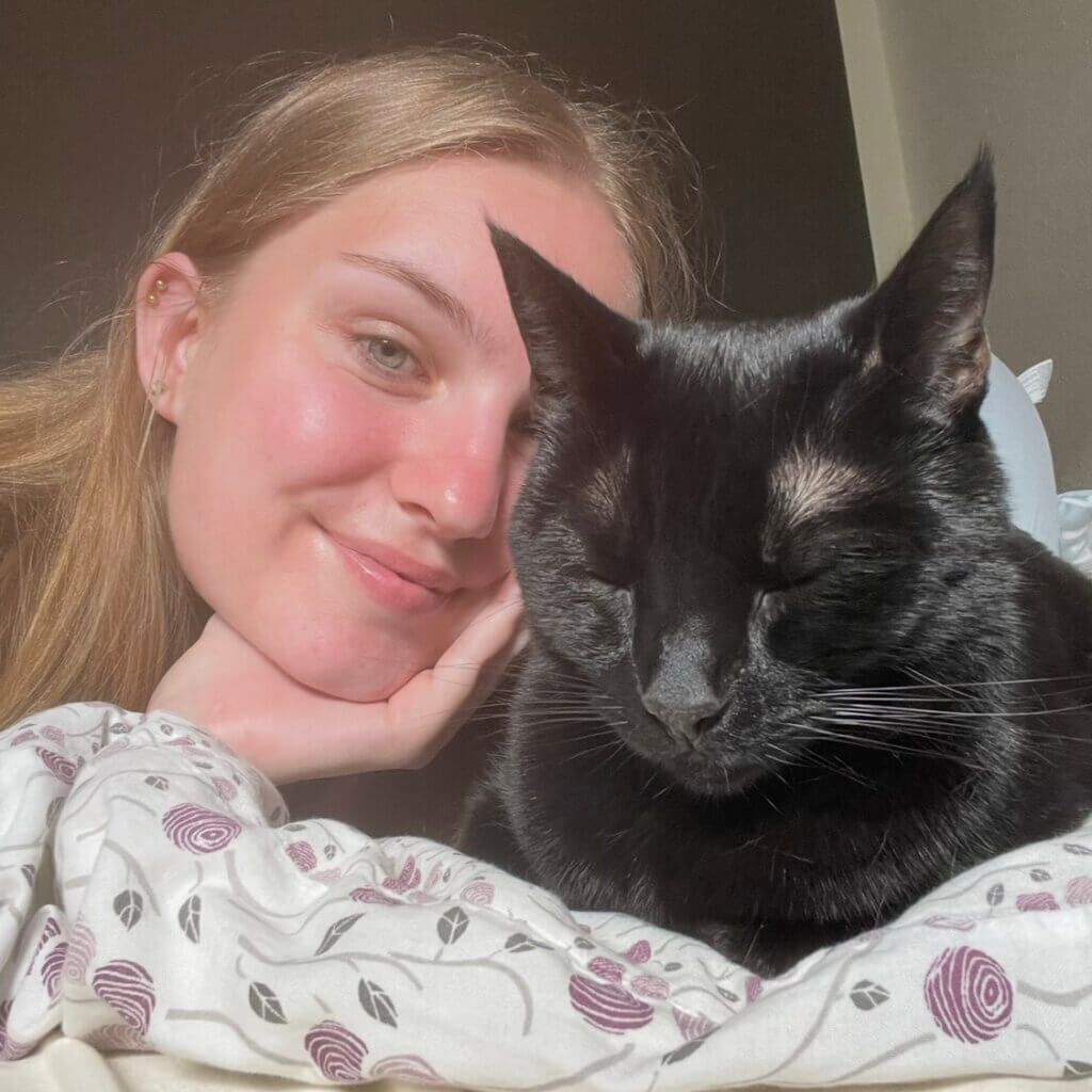 A black cat sits atop a bed with Nathalie Nielsen's face in the frame.