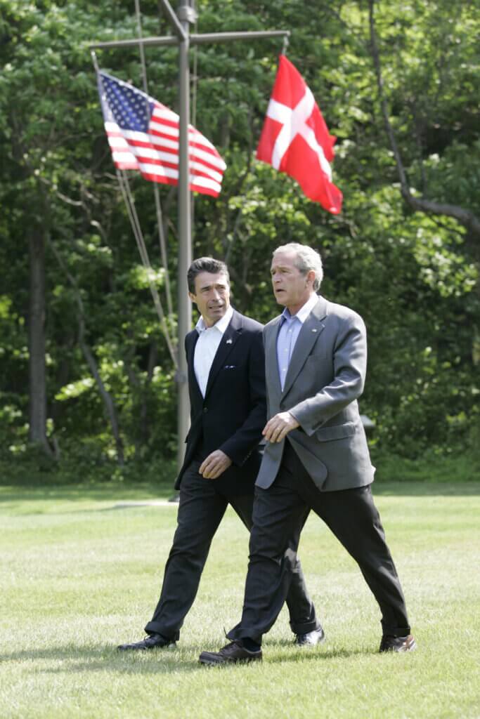 Two men are walking on a lawn, they are both wearing suits, behind them are the Danish and American flags hanging.