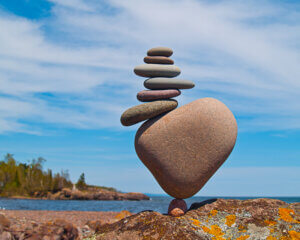 A rock cairn, a small rock has a big rock balancing on it, and on top of the big rock are several smaller flat rocks that are balancing on top of each other. Behind is a blue lake and sky