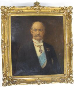 Portrait of an older man in a dark suit with medals and a blue sash, set against a brown background and a gold frame.