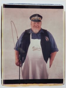 Photograph of a man wearing a white apron, black vest, blue shirt, and a cap. He is holding a whip in his right hand.
