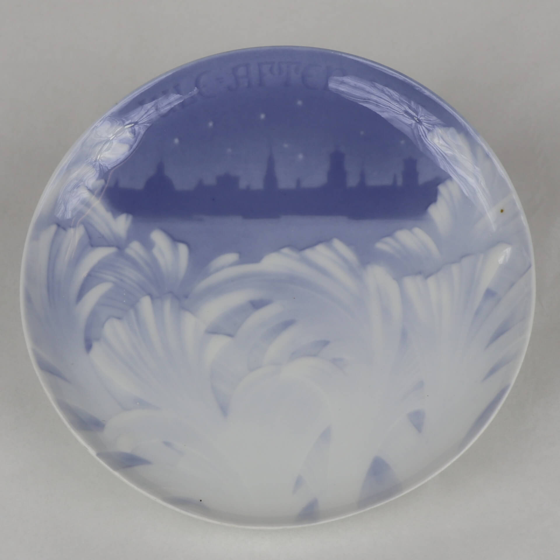 A ceramic plate in blue and white. A leaf-like motif is in the bottom two-thirds. in the third is the silhouette of a city. Text at the top reads "Jule Aften 1895."