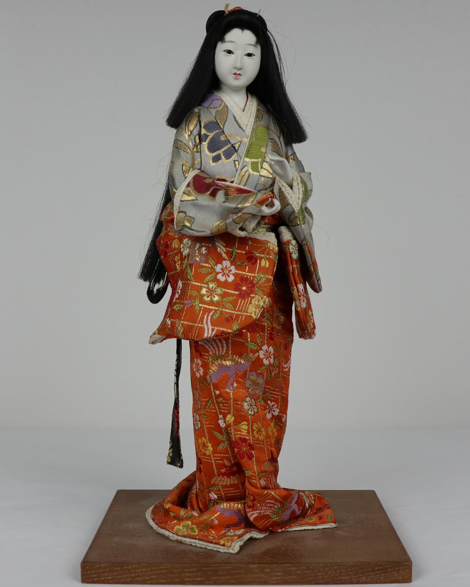 A Japanese doll standing on a wooden base, wearing an orange and gray kimono with floral pattern. It has long black hair.