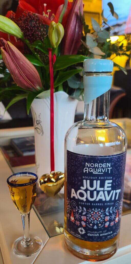 The 2024 Jule Aquavit release. A small glass for aquavit is filled with amber liquid. A bottle with a blue label is placed beside the glass. The bottle is partially full.