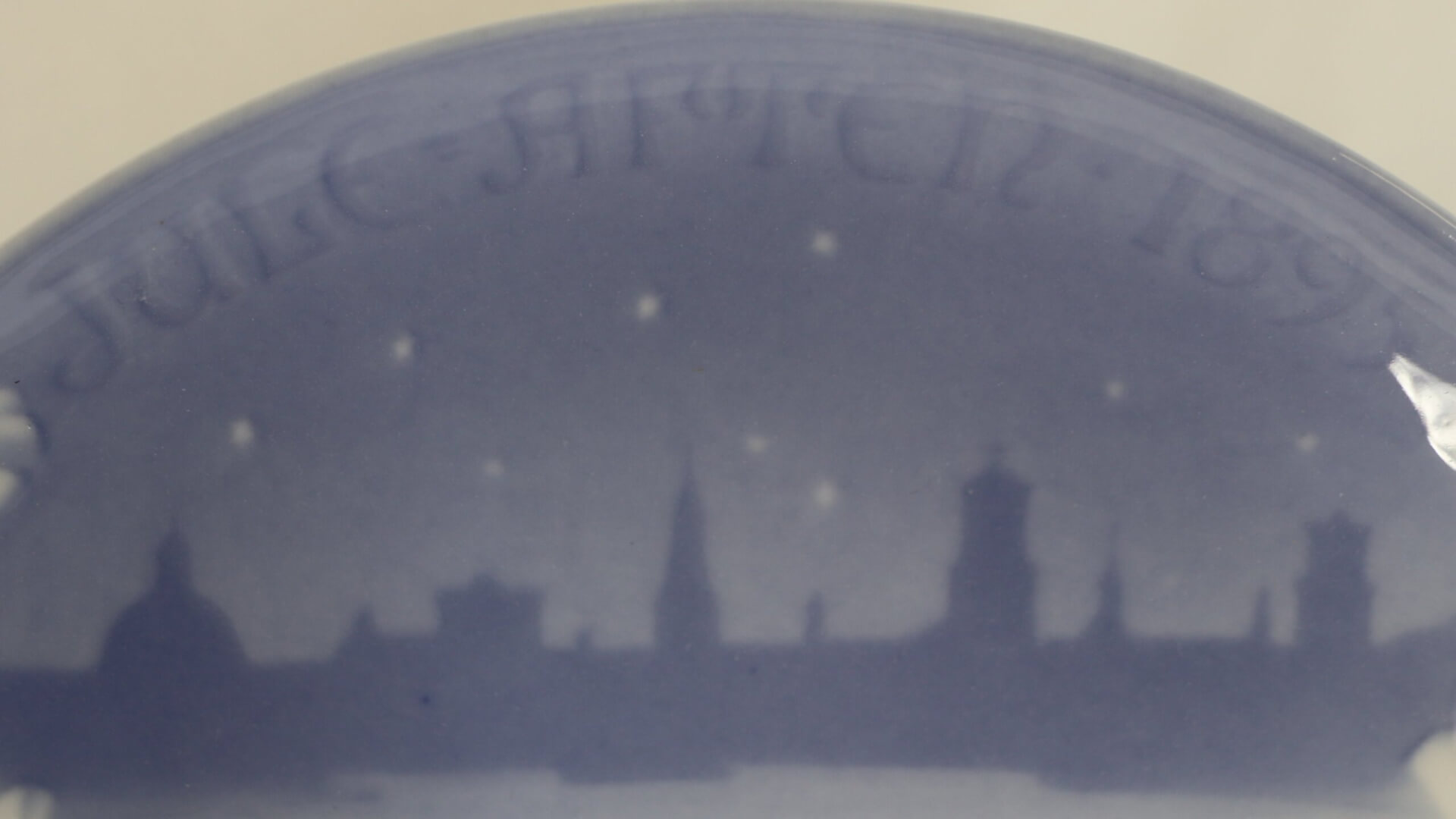 A ceramic plate in blue and white. The top has text reading "Jule Aften 1895." The bottom has a silhouette of a city.