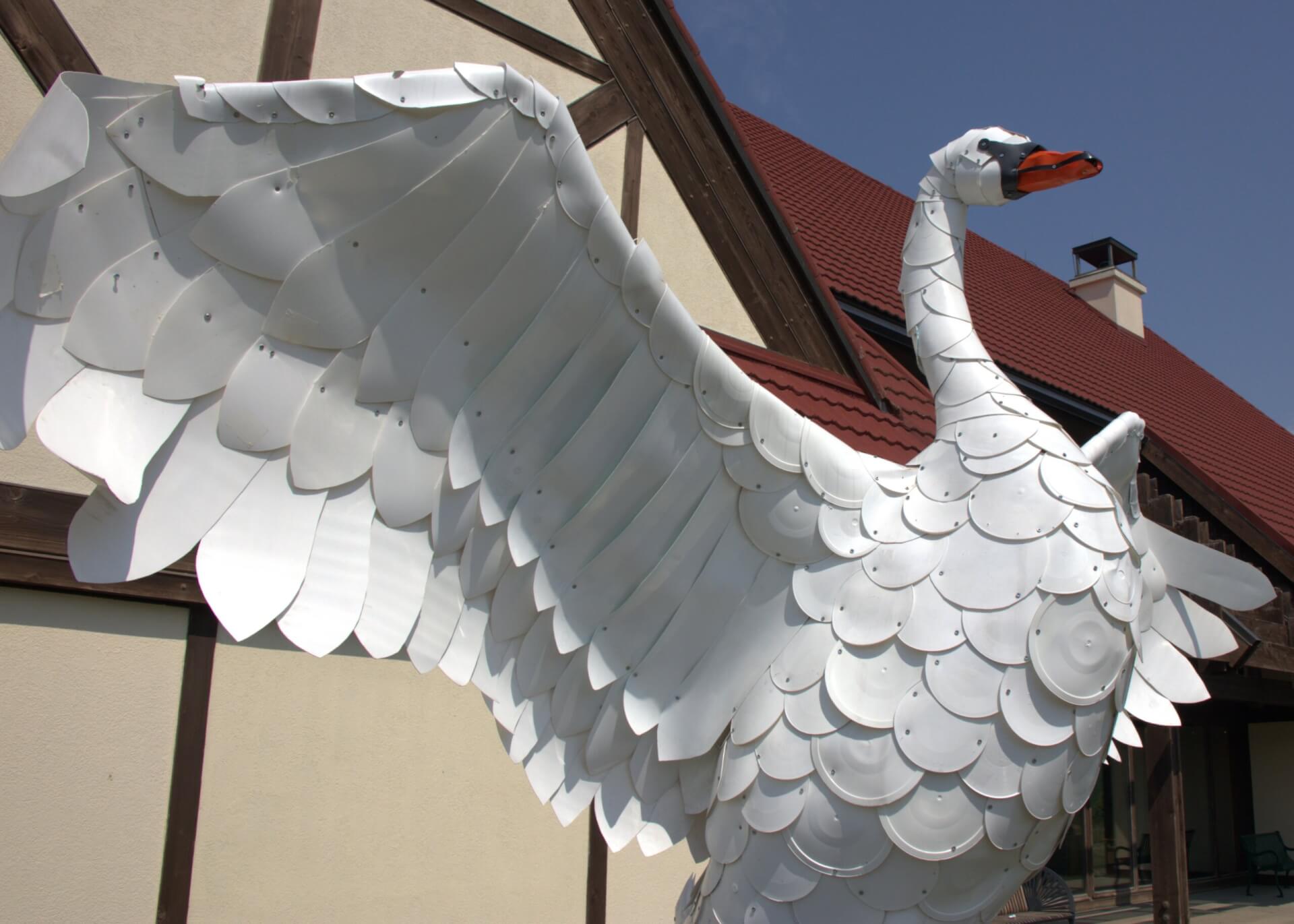 Swan sculpture made of white plastic. Blue sky and a white building with a red roof are behind it.