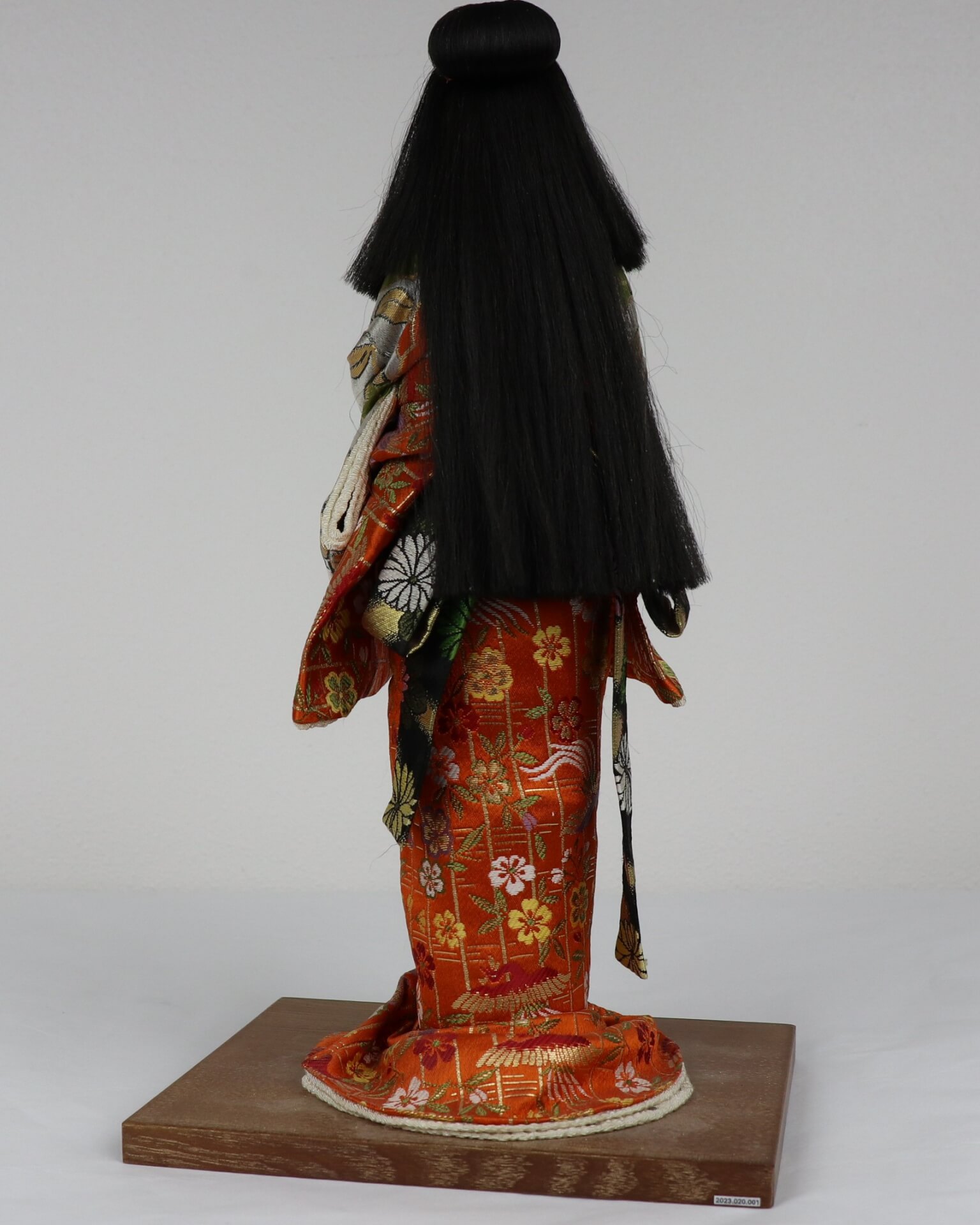 Backside of Japanese doll with long black hair and an orange kimono.