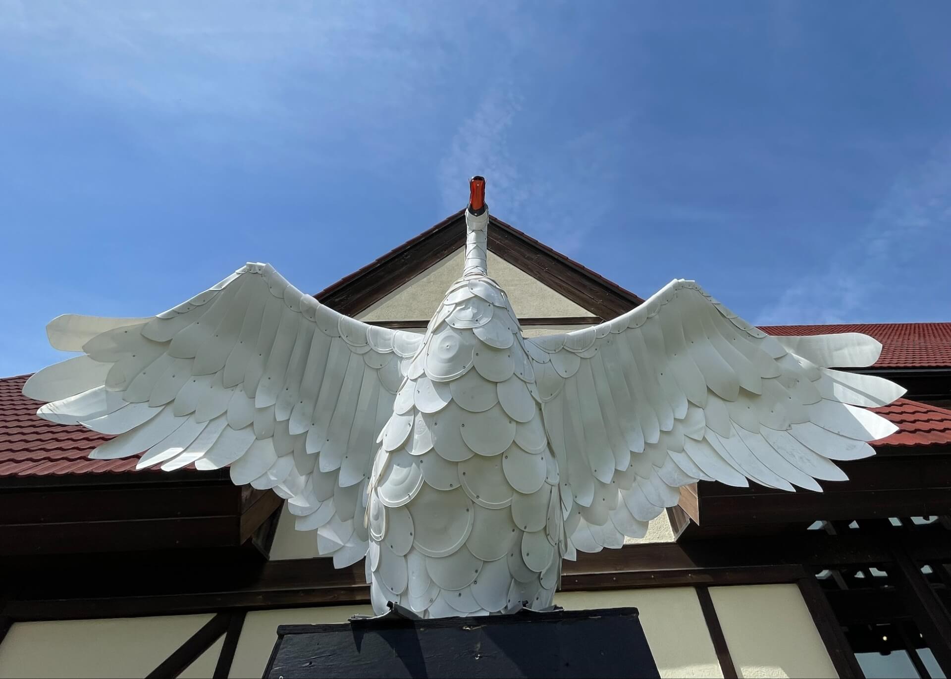 Swan sculpture made of white plastic. Blue sky and a white building with a red roof is behind it.