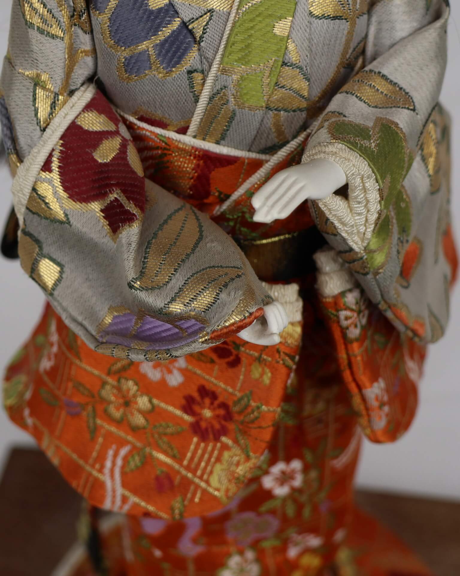 Close up of hands and pattern of kimono. Gold and green leaves dominate the gray of the kimono, multiple colored flowers dominate the orange part.