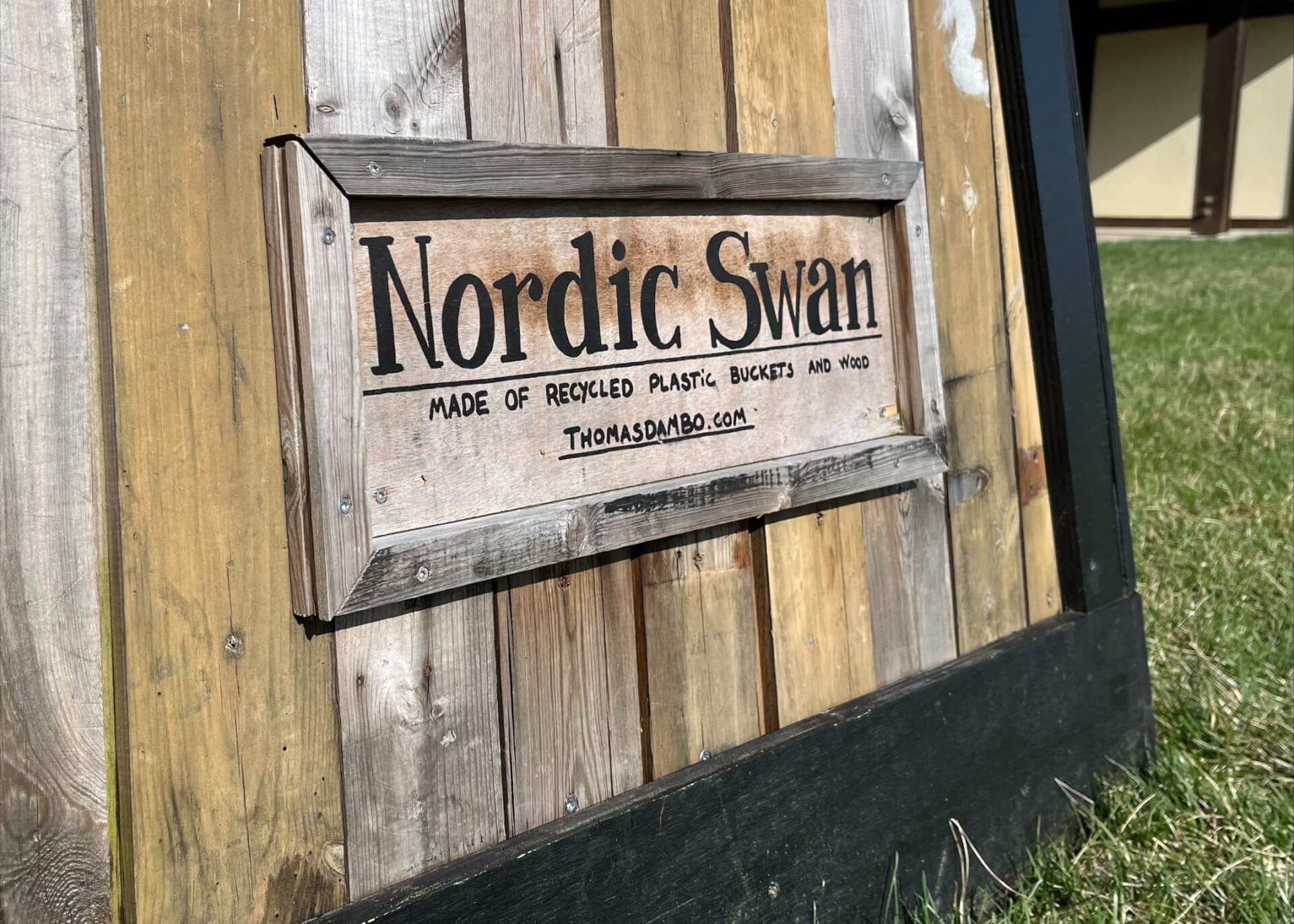 Wooden sign attached to a rustic wooden crate. Text on the sign reads "Nordic Swan. Made of Recycled Plastic Buckets and wood. Thomasdambo.com."