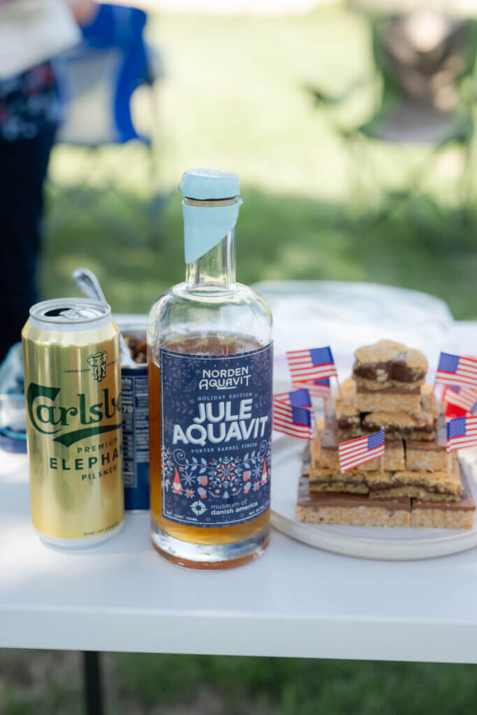 A gold can of Carlsberg, an opened bottle of Jule Aquavit, and Midwestern bars decorated with American flags on a folding table.