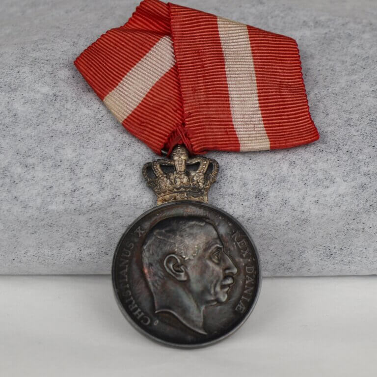 A tarnished silver medal with a red and white striped ribbon. The medal features a man in profile, with text reading "Christianus X Rex Daniae." On top of the medal is a crown.