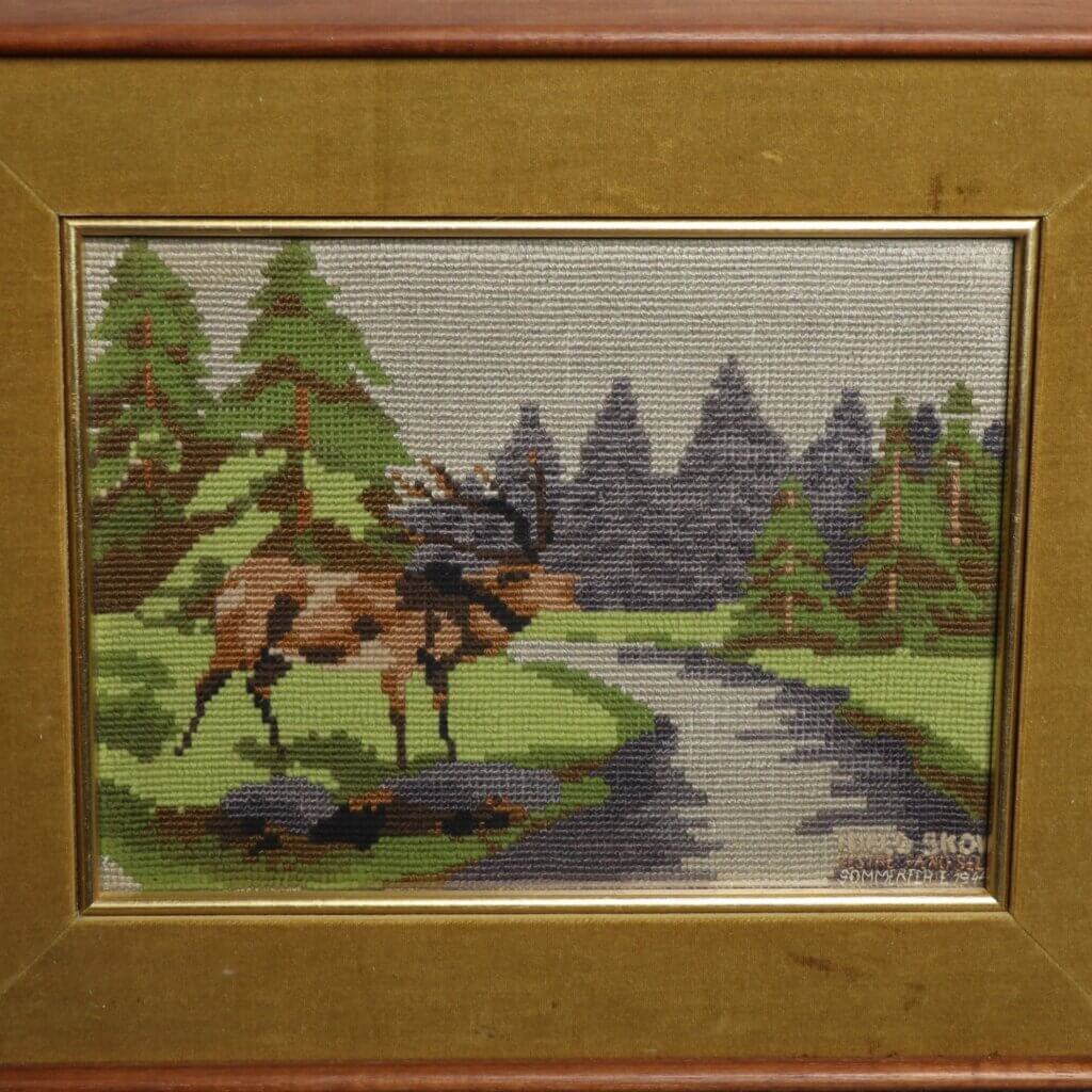 Framed cross stitch of a red deer near a stream, surrounded by green trees.