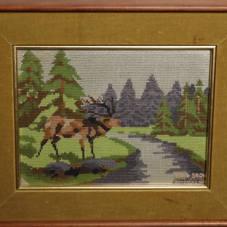 Framed cross stitch of a red deer near a stream, surrounded by green trees.