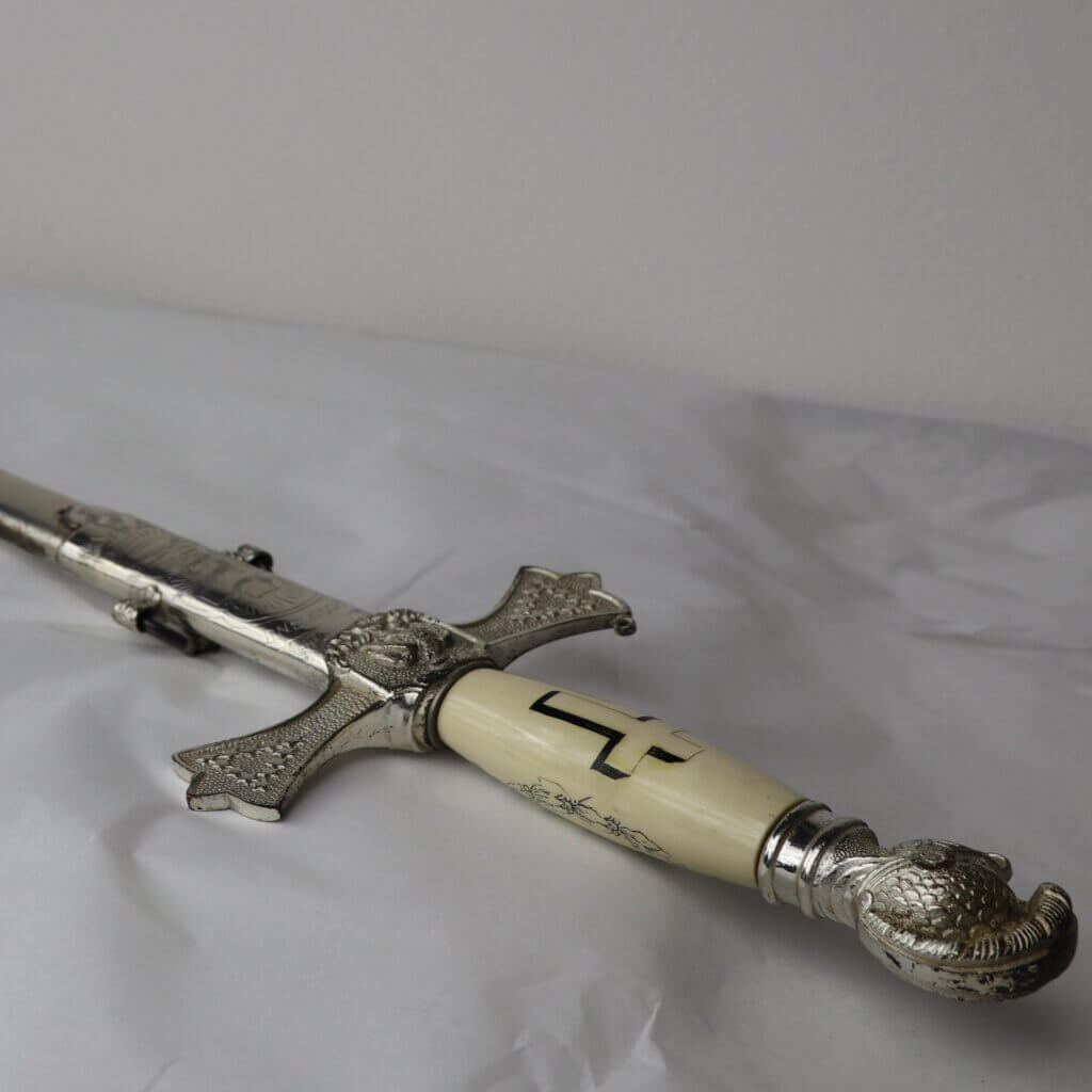 Ceremonial sword in ornate scabbard, featuring engravings and an ivory colored handle with a cross.
