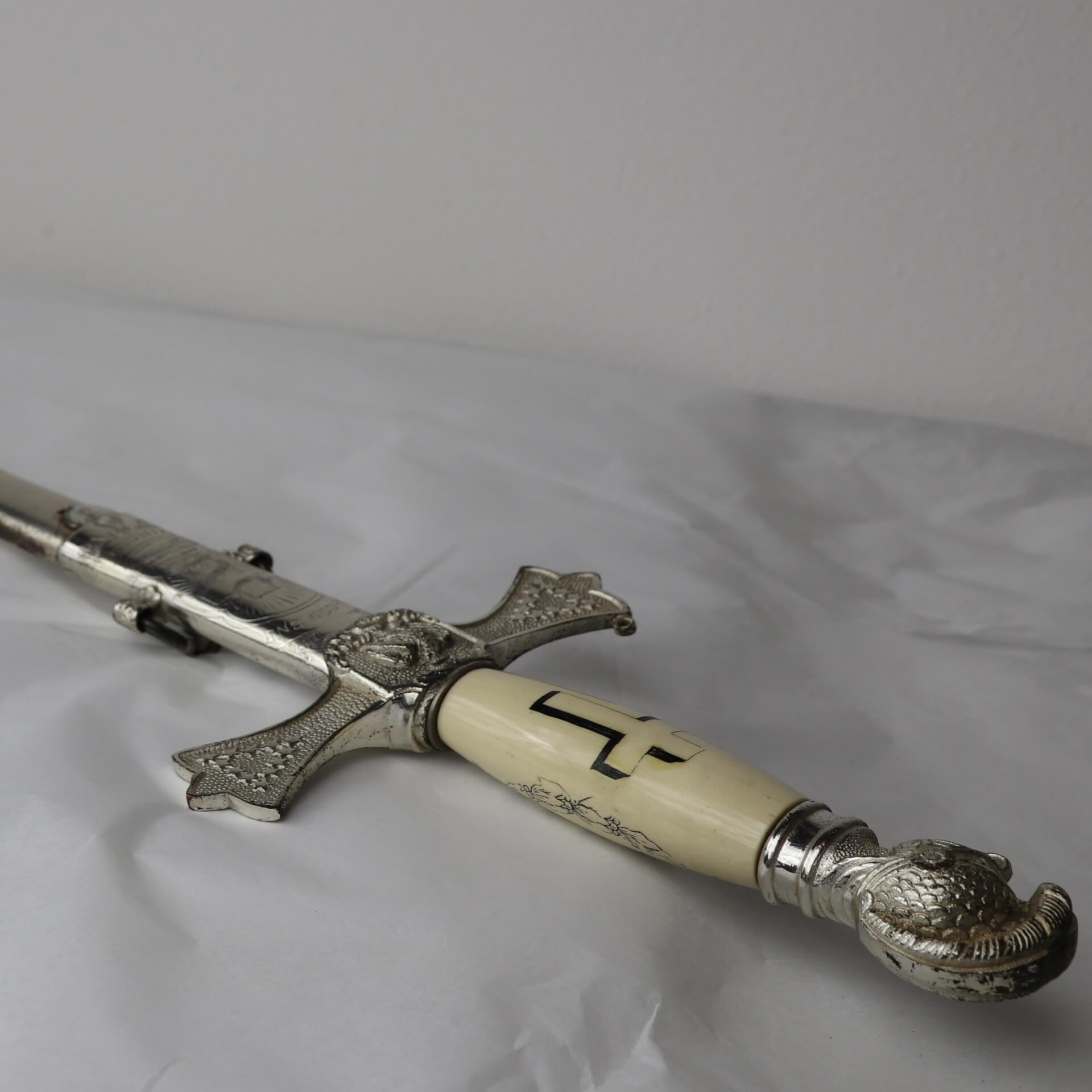 Ceremonial sword in ornate scabbard, featuring engravings and an ivory colored handle with a cross.