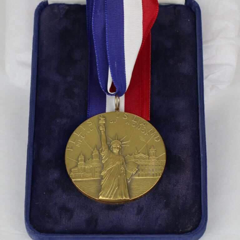 A Brass medal with the Statue of Liberty. It hangs on a red, white, and blue ribbon. Text on the rim of the medal reads "Ellis Island Medal of Honor."