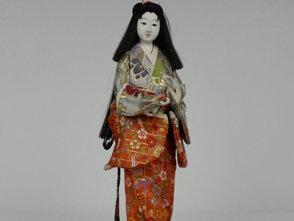 A Japanese doll standing on a wooden base, wearing an orange and gray kimono with floral pattern. It has long black hair.