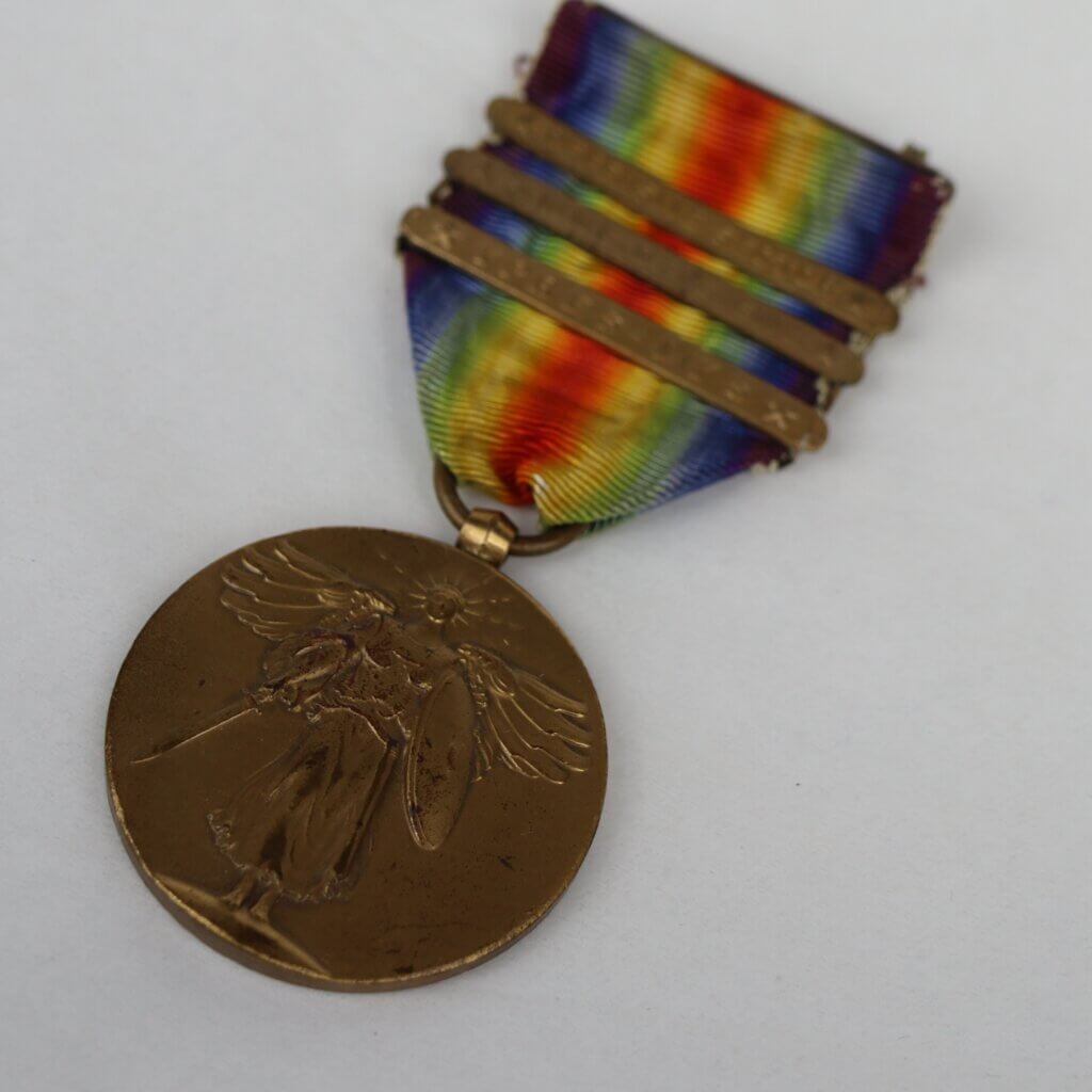 A bronze medal hangs from a rainbow striped ribbon with three bronze clasps. The medal has a winged woman holding a sword and shield.