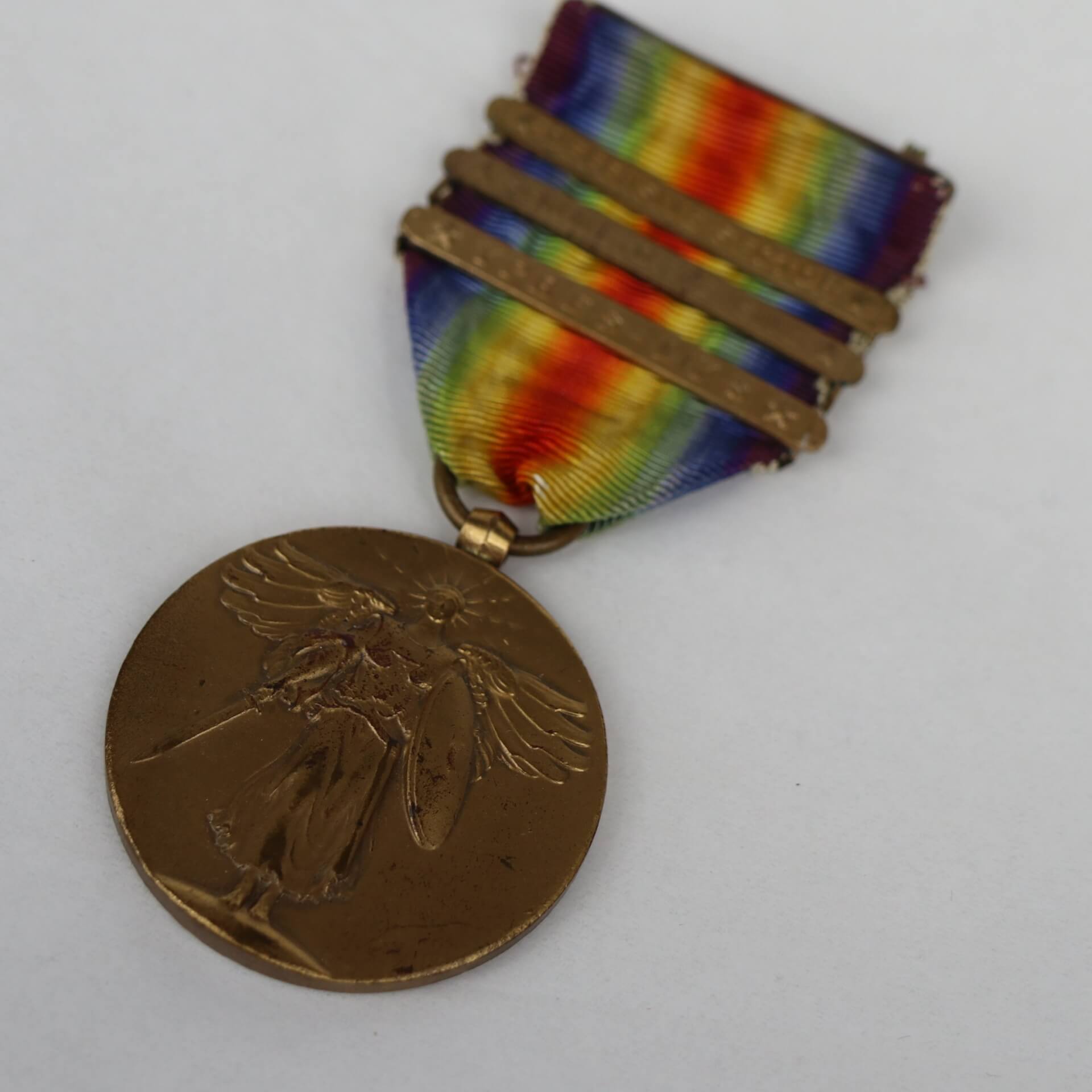 A bronze medal hangs from a rainbow striped ribbon with three bronze clasps. The medal has a winged woman holding a sword and shield.