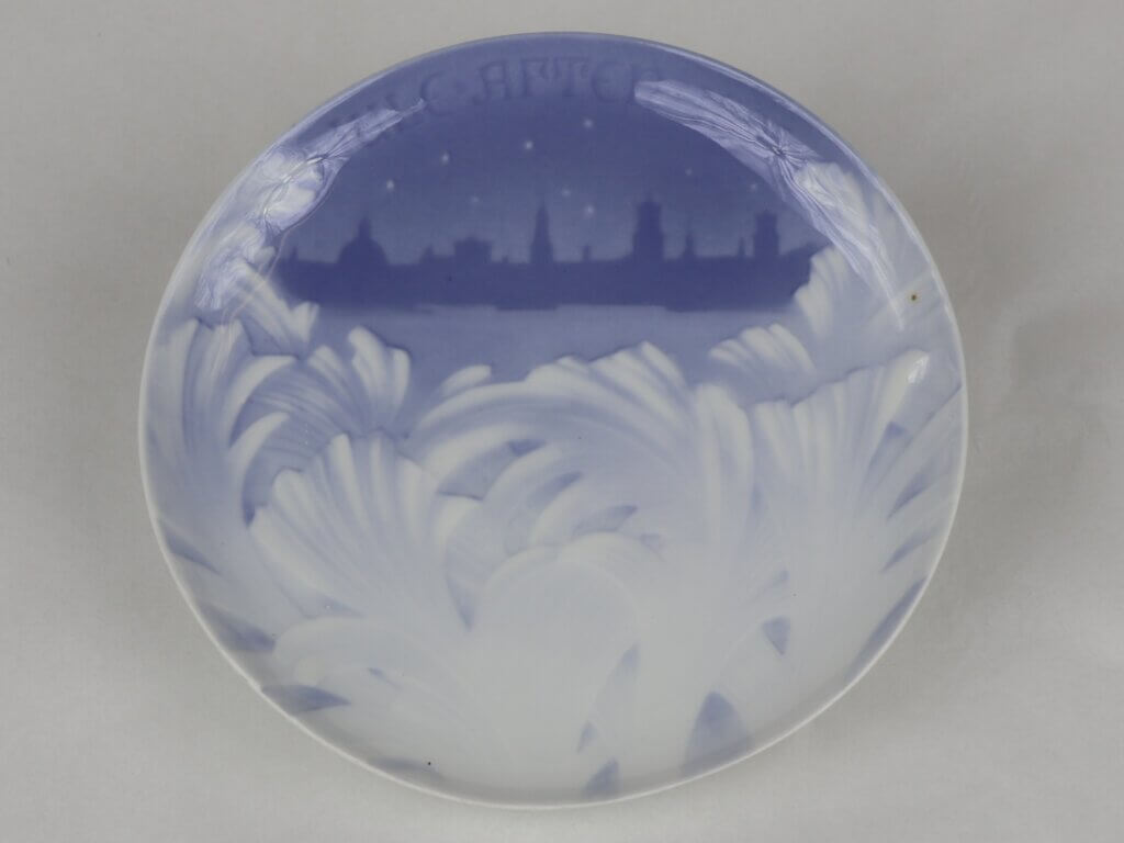 A ceramic plate in blue and white. A leaf-like motif occurs on the bottom of the plate, while the top third shows the silhouette of a city. Text at the top reads "Jule Aften 1895"