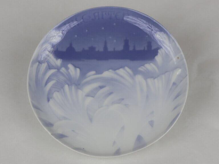 A ceramic plate in blue and white. A leaf-like motif occurs on the bottom of the plate, while the top third shows the silhouette of a city. Text at the top reads "Jule Aften 1895"