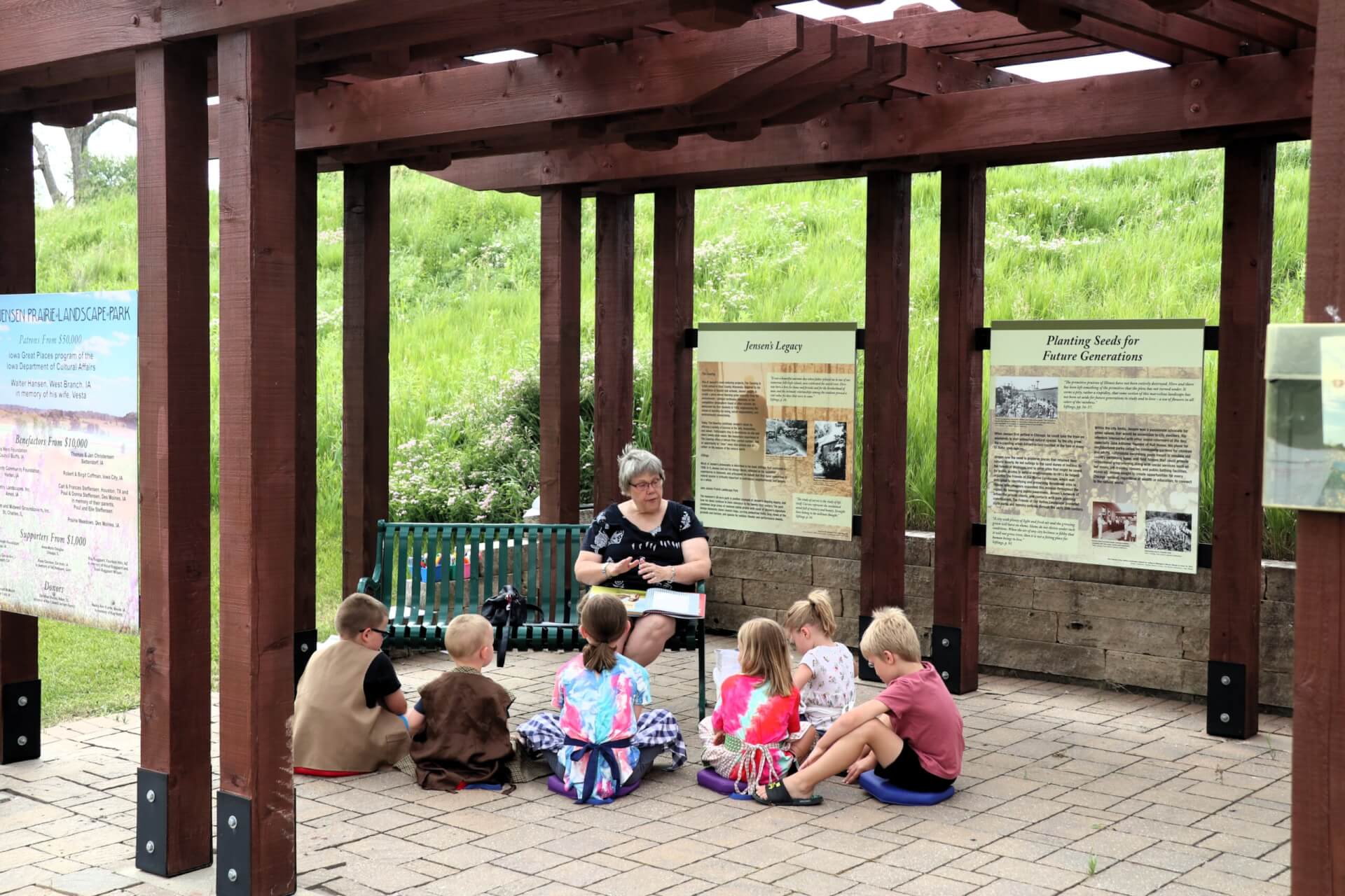 A woman sits on a green bench reading books to children under a dark, wooden pergola.