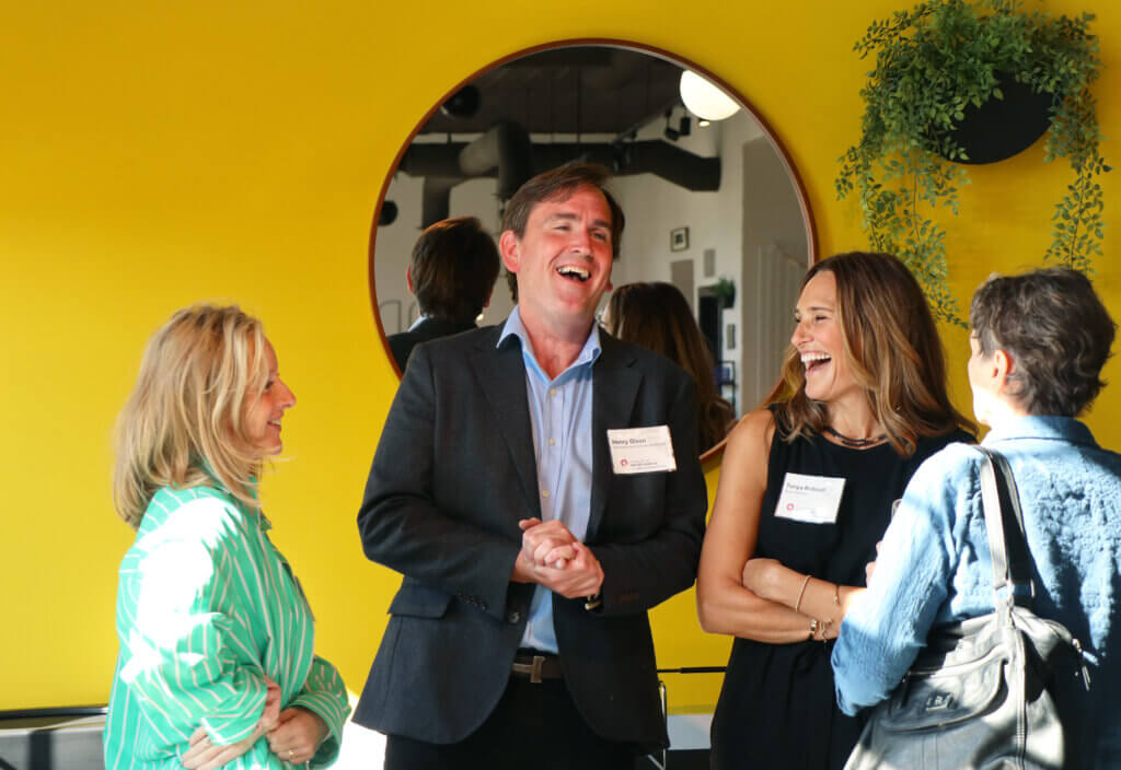 A MoDA staff member laughs with board members and a Danish intern before a mirror and hanging plant.