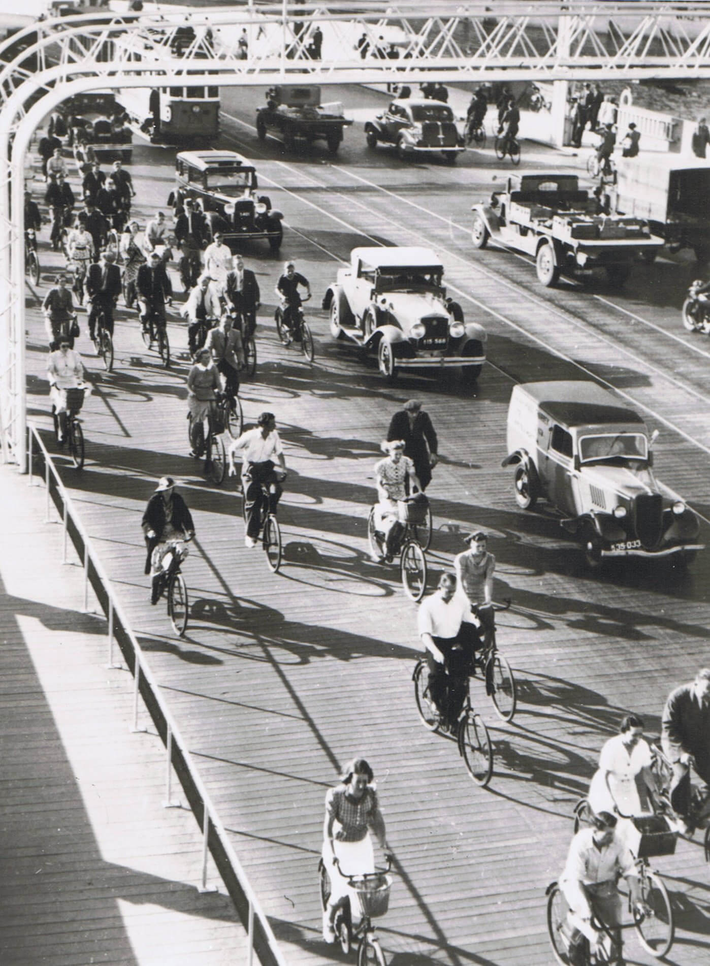 Black and White photograph with old cars on the right and a line of bicycling people on the left.