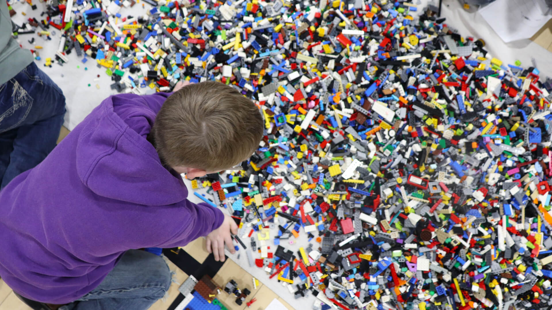 A young boy wearing a purple sweatshirt and blue jeans kneels before a floor filled with LEGOs.
