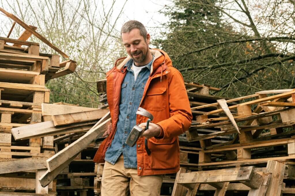 Thomas Dambo, wearing an orange jacket and blue button up, posing with pallets and tools.