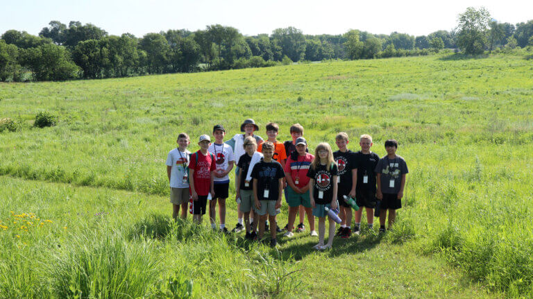 A group of children wearing lanyards and holding water bottles stand on a mown path in the Jens Jensen Prairie Landscape Park.